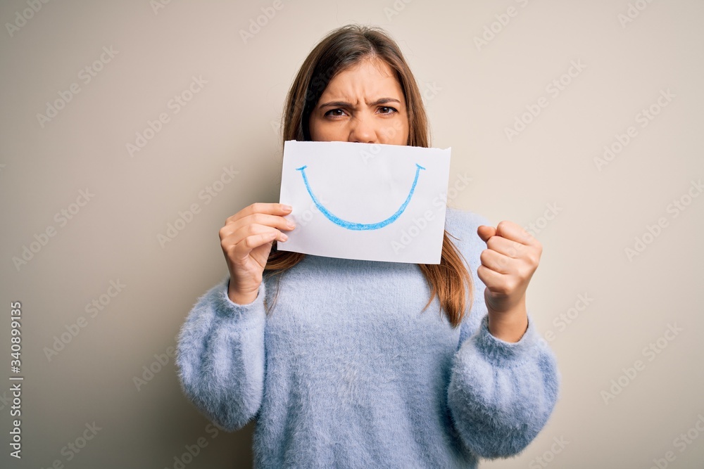 Young blonde woman holding funny smile drawing on mouth as happy expression annoyed and frustrated shouting with anger, crazy and yelling with raised hand, anger concept Young blonde woman holding funny smile drawing on mouth as happy expression annoyed and frustrated shouting with anger, crazy and yelling with raised hand, anger concept