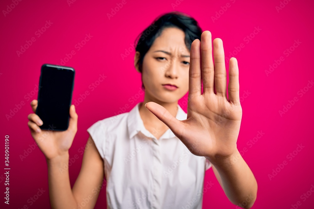 Young beautiful asian girl holding smartphone showing screen over isolated pink background with open hand doing stop sign with serious and confident expression, defense gesture Young beautiful asian girl holding smartphone showing screen over isolated pink background with open hand doing stop sign with serious and confident expression, defense gesture