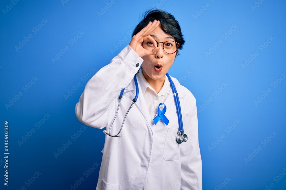 Young beautiful asian doctor girl wearing stethoscope and coat with blue cancer ribbon doing ok gesture shocked with surprised face, eye looking through fingers. Unbelieving expression. Young beautiful asian doctor girl wearing stethoscope and coat with blue cancer ribbon doing ok gesture shocked with surprised face, eye looking through fingers. Unbelieving expression.