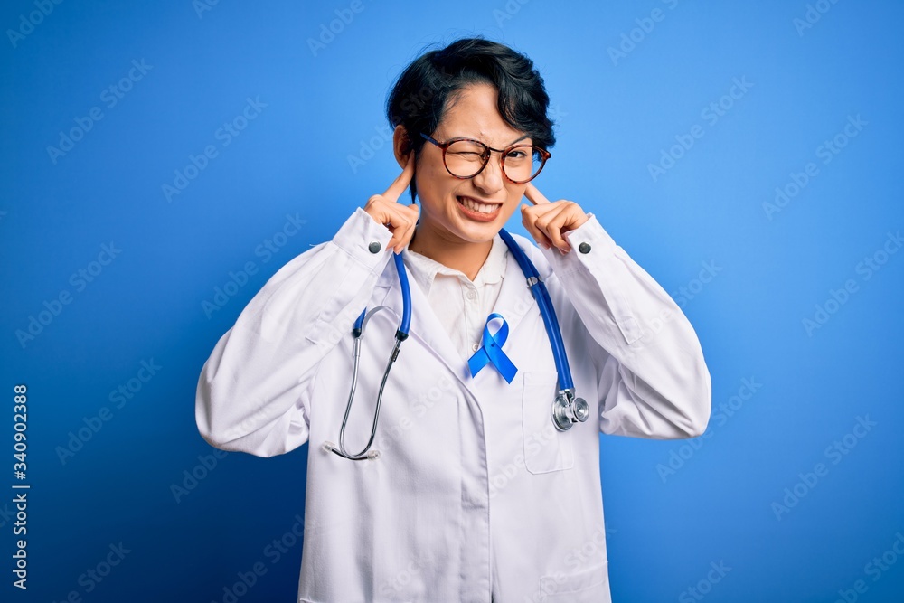 Young beautiful asian doctor girl wearing stethoscope and coat with blue cancer ribbon covering ears with fingers with annoyed expression for the noise of loud music. Deaf concept. Young beautiful asian doctor girl wearing stethoscope and coat with blue cancer ribbon covering ears with fingers with annoyed expression for the noise of loud music. Deaf concept.