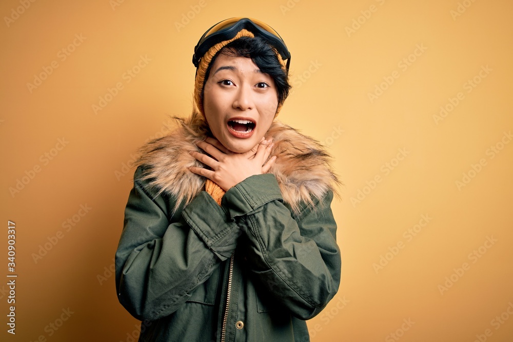 Beautiful asian skier girl wearing snow sportswear using ski goggles over yellow background shouting and suffocate because painful strangle. Health problem. Asphyxiate and suicide concept.