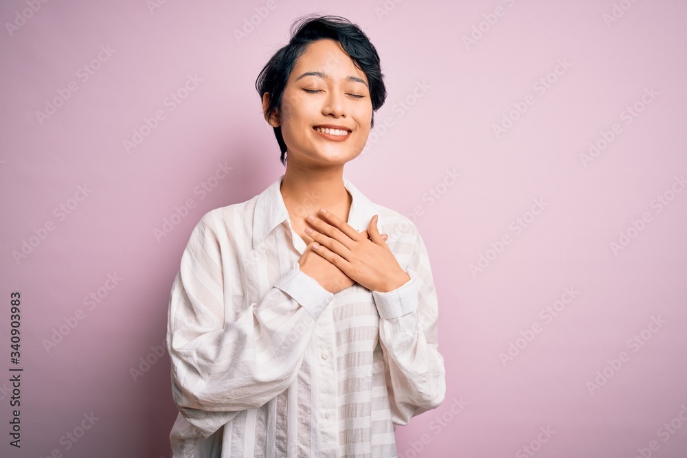 Young beautiful asian girl wearing casual shirt standing over isolated pink background smiling with hands on chest with closed eyes and grateful gesture on face. Health concept. Young beautiful asian girl wearing casual shirt standing over isolated pink background smiling with hands on chest with closed eyes and grateful gesture on face. Health concept.