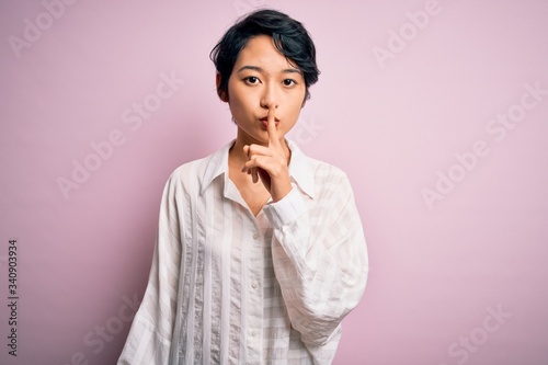 Young beautiful asian girl wearing casual shirt standing over isolated pink background asking to be quiet with finger on lips. Silence and secret concept.