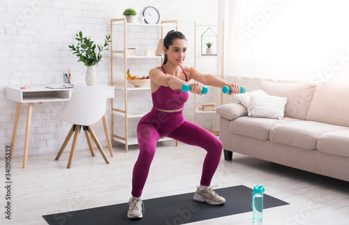 Fototapeta Naklejka Na Ścianę i Meble -  At home sports. Sporty girl doing squats with dumbbells in living room