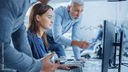 Modern Factory Office: Project Manager and Supervisor Talk to a Female Engineer Works on Computer. Team of Professionals Solving Heavy Industry Problems, Having Discussion, Pointing at Display