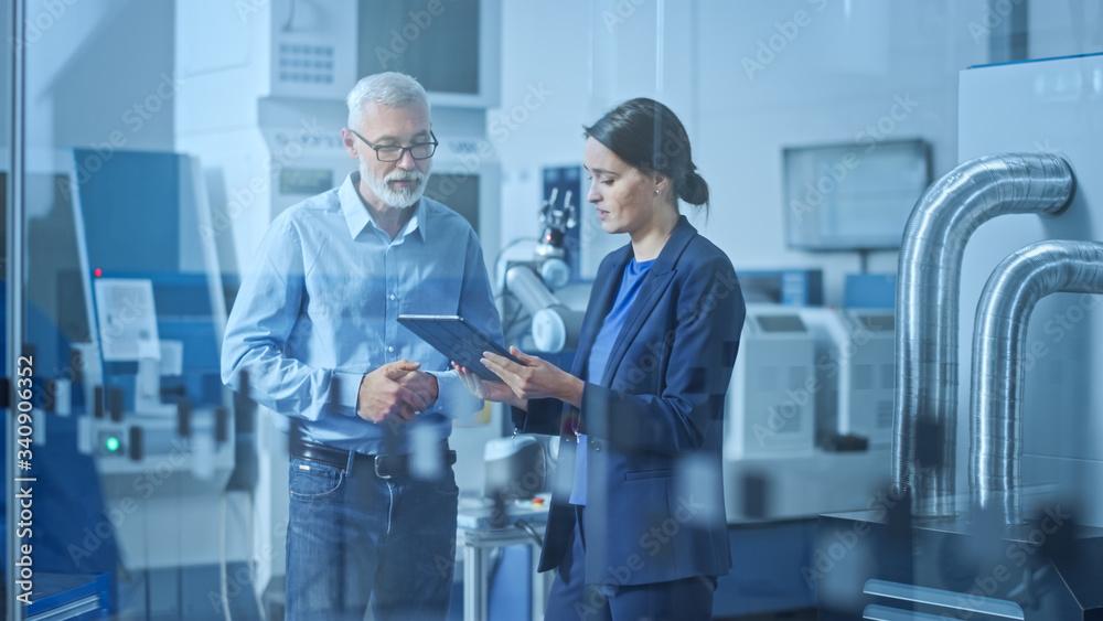 Modern Factory: Female Engineer, Male Project Manager Standing in High Tech Development Facility, Talking and Using Tablet Computer. Contemporary Facility with CNC Machinery, Robot Arm Production Line Modern Factory: Female Engineer, Male Project Manager Standing in High Tech Development Facility, Talking and Using Tablet Computer. Contemporary Facility with CNC Machinery, Robot Arm Production Line