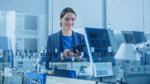Modern Factory Office: Portrait of Young and Confident Female Industrial Engineer Standing and Holding Digital Tablet. Industrial Factory Office with Glass Wall and CNC Machinery Workshop Behind