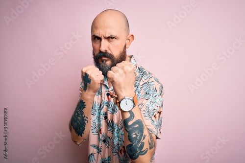 Handsome bald man with beard and tattoo wearing casual floral shirt over pink background Ready to fight with fist defense gesture, angry and upset face, afraid of problem