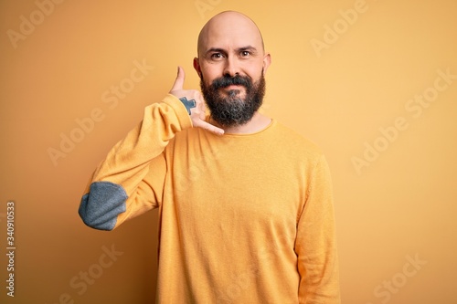 Handsome bald man with beard wearing casual sweater standing over yellow background smiling doing phone gesture with hand and fingers like talking on the telephone. Communicating concepts.