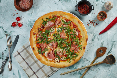 Freshly baked crispy pizza with prosciutto crudo, parmesan, mushrooms and arugula on a gray background in a composition with ingredients and kitchen utensils