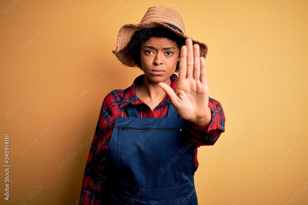 Young African American afro farmer woman with curly hair wearing apron and hat doing stop sing with palm of the hand. Warning expression with negative and serious gesture on the face. Young African American afro farmer woman with curly hair wearing apron and hat doing stop sing with palm of the hand. Warning expression with negative and serious gesture on the face.