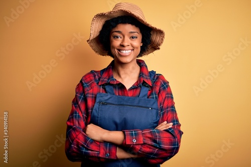 Young African American afro farmer woman with curly hair wearing apron and hat happy face smiling with crossed arms looking at the camera. Positive person.