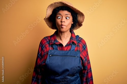 Young African American afro farmer woman with curly hair wearing apron and hat making fish face with lips, crazy and comical gesture. Funny expression.