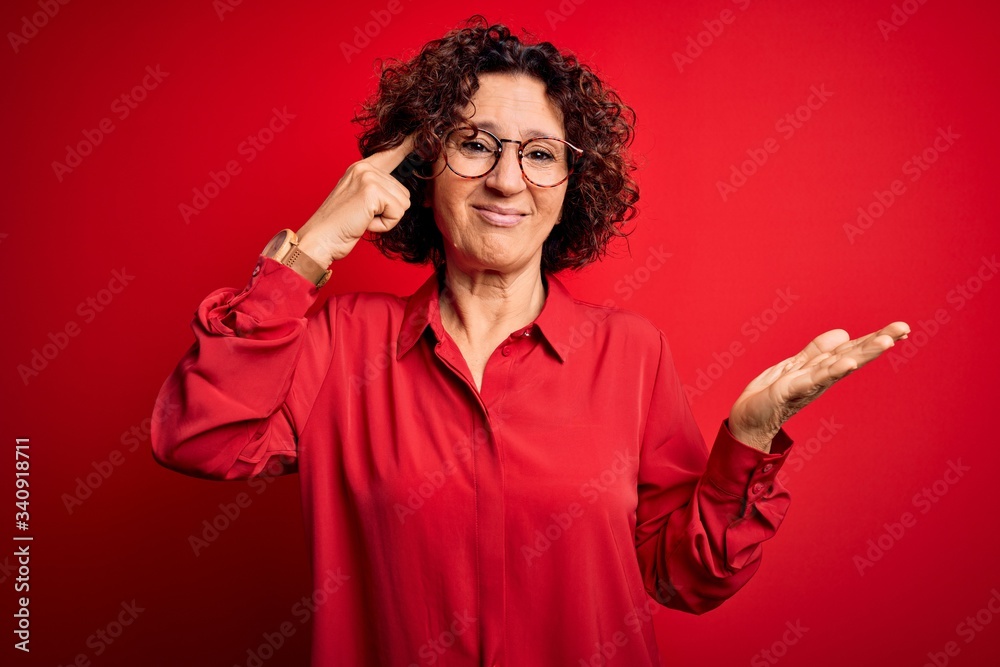 Middle age beautiful curly hair woman wearing casual shirt and glasses over red background confused and annoyed with open palm showing copy space and pointing finger to forehead. Think about it. Middle age beautiful curly hair woman wearing casual shirt and glasses over red background confused and annoyed with open palm showing copy space and pointing finger to forehead. Think about it.