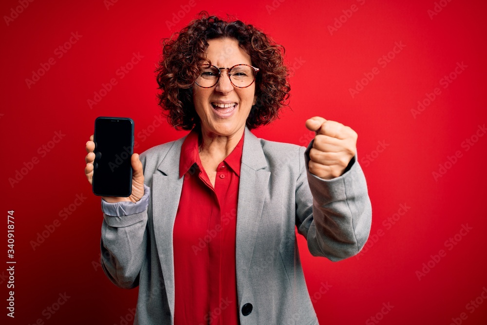 Middle age curly woman holding smartphone showing screen over isolated red background screaming proud and celebrating victory and success very excited, cheering emotion Middle age curly woman holding smartphone showing screen over isolated red background screaming proud and celebrating victory and success very excited, cheering emotion