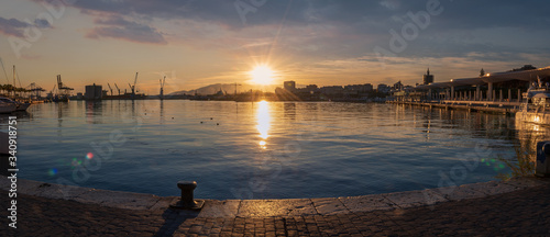 Sunset in Malaga Port wide panorama from Quay 1 in Spain with sun reflections in the sea water and cranes.