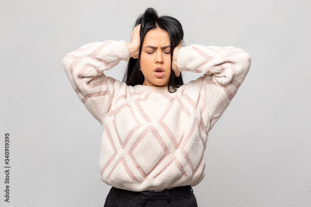 Portrait of a beautiful brunette in a warm white winter sweater. The young woman has a headache, on a gray background. The girl holds her hands behind her head Portrait of a beautiful brunette in a warm white winter sweater. The young woman has a headache, on a gray background. The girl holds her hands behind her head