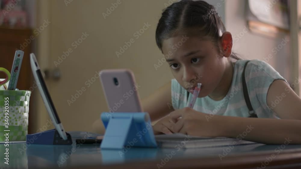Adorable asian girl watching lesson online from mobile phone and digital tablet during doing homework. Female teenager studying from home with video call during COVID-19 pandemic situation. Adorable asian girl watching lesson online from mobile phone and digital tablet during doing homework. Female teenager studying from home with video call during COVID-19 pandemic situation.
