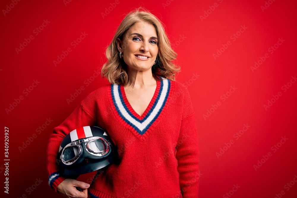 Fototapeta premium Middle age beautiful blonde motorcyclist woman holding moto helmet over red background with a happy face standing and smiling with a confident smile showing teeth