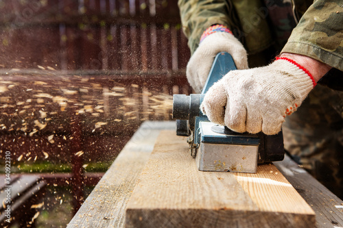 Hands of a joiner with an electric planer. Flying shavings, wooden board. Woodwork. The work of a professional.