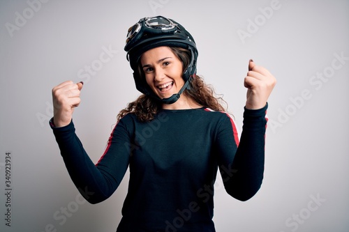 Beautiful motorcyclist woman with curly hair wearing moto helmet over white background celebrating surprised and amazed for success with arms raised and open eyes. Winner concept.