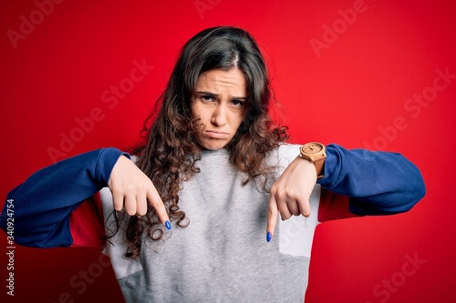 Young beautiful woman with curly hair wearing casual sweatshirt over isolated red background Pointing down looking sad and upset, indicating direction with fingers, unhappy and depressed. © Krakenimages.com