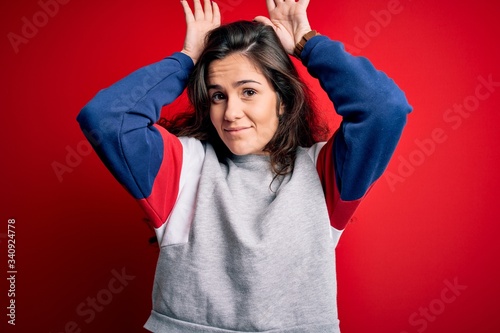 Young beautiful woman with curly hair wearing casual sweatshirt over isolated red background Doing bunny ears gesture with hands palms looking cynical and skeptical. Easter rabbit concept. photo