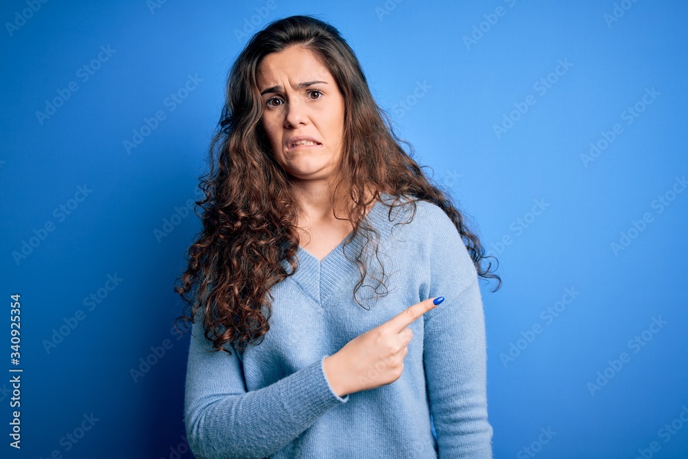 Young beautiful woman with curly hair wearing blue casual sweater over isolated background Pointing aside worried and nervous with forefinger, concerned and surprised expression Young beautiful woman with curly hair wearing blue casual sweater over isolated background Pointing aside worried and nervous with forefinger, concerned and surprised expression
