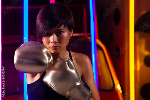 Athlete Asian Woman is training punching Silver Mitts Gloves Boxer. Office Girl exercises in Modern color Neon "Muay Thai Tuk Tuk car" Boxing Gym background, low exposure high saturation