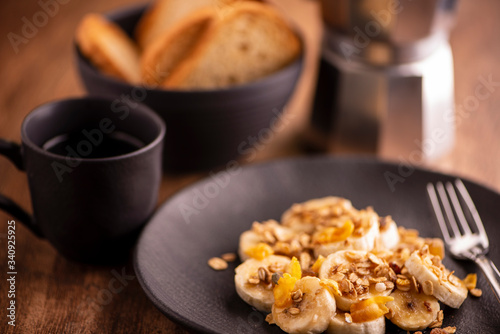 plate with sliced banana with granola, candied fruit and honey, toast and a cup of coffee