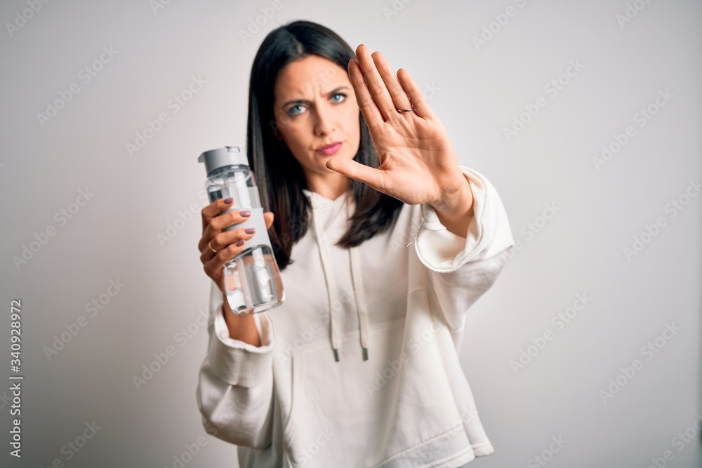 Young brunette sporty woman with blue eyes holding water bottle over white background with open hand doing stop sign with serious and confident expression, defense gesture Young brunette sporty woman with blue eyes holding water bottle over white background with open hand doing stop sign with serious and confident expression, defense gesture