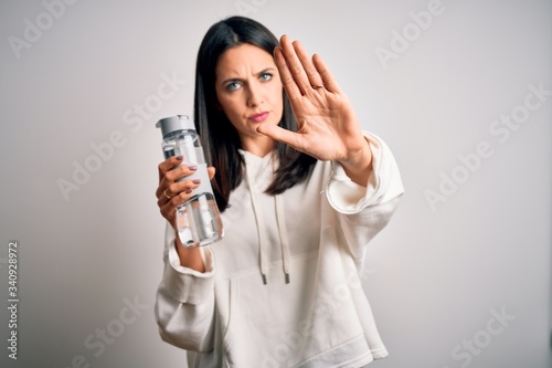 Young brunette sporty woman with blue eyes holding water bottle over white background with open hand doing stop sign with serious and confident expression, defense gesture