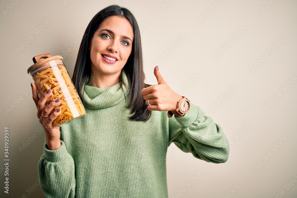 Young brunette woman holding dry italian macaroni over brown isolated background happy with big smile doing ok sign, thumb up with fingers, excellent sign Young brunette woman holding dry italian macaroni over brown isolated background happy with big smile doing ok sign, thumb up with fingers, excellent sign