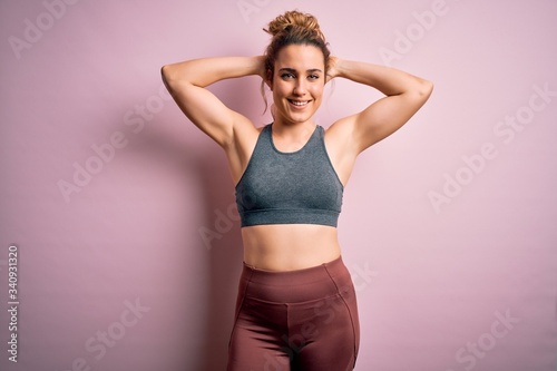 Young beautiful blonde sportswoman doing sport wearing sportswear over pink background relaxing and stretching, arms and hands behind head and neck smiling happy © Krakenimages.com