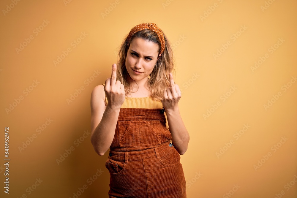 Young beautiful blonde woman wearing overalls and diadem standing over yellow background Showing middle finger doing fuck you bad expression, provocation and rude attitude. Screaming excited Young beautiful blonde woman wearing overalls and diadem standing over yellow background Showing middle finger doing fuck you bad expression, provocation and rude attitude. Screaming excited