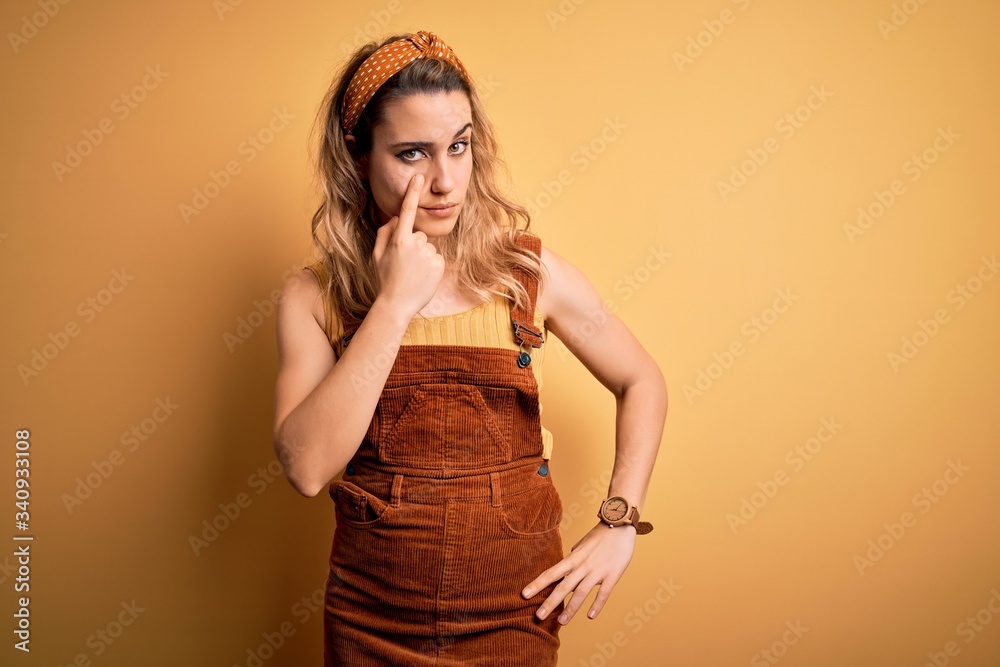 Young beautiful blonde woman wearing overalls and diadem standing over yellow background Pointing to the eye watching you gesture, suspicious expression Young beautiful blonde woman wearing overalls and diadem standing over yellow background Pointing to the eye watching you gesture, suspicious expression