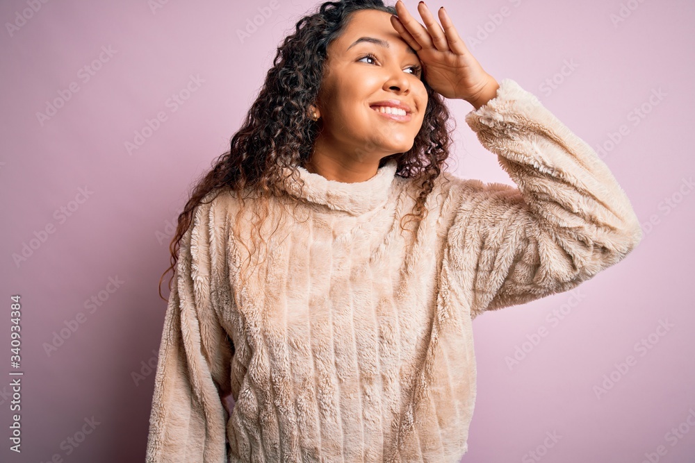 Young beautiful woman with curly hair wearing casual sweater standing over pink background very happy and smiling looking far away with hand over head. Searching concept. Young beautiful woman with curly hair wearing casual sweater standing over pink background very happy and smiling looking far away with hand over head. Searching concept.