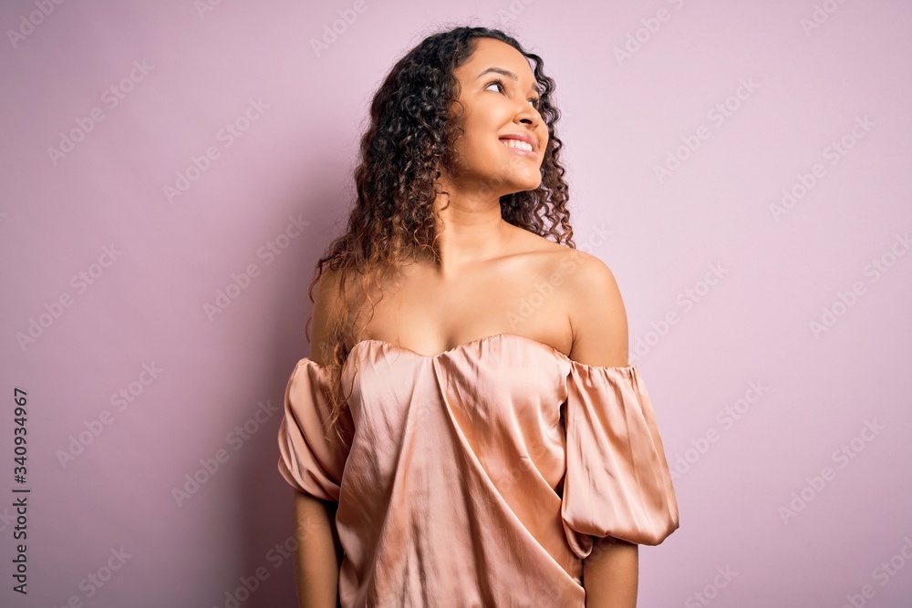 Young beautiful woman with curly hair wearing casual t-shirt standing over pink background looking away to side with smile on face, natural expression. Laughing confident. Young beautiful woman with curly hair wearing casual t-shirt standing over pink background looking away to side with smile on face, natural expression. Laughing confident.