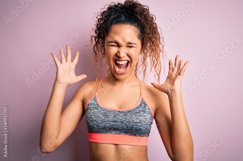 Beautiful sportswoman with curly hair doing sport wearing sportswear over pink background celebrating mad and crazy for success with arms raised and closed eyes screaming excited. Winner concept