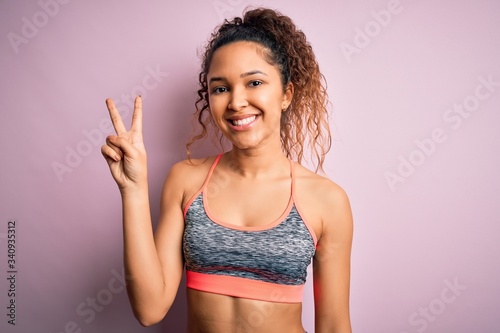Beautiful sportswoman with curly hair doing sport wearing sportswear over pink background showing and pointing up with fingers number two while smiling confident and happy. © Krakenimages.com