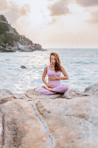 Pregnant woman practices yoga  sitting in lotus position on the beach  on the rocks. Hands of girl in a pose meditating. Serenity practicing at sunrise  meditation. Woman in a pink-purple tracksuit.