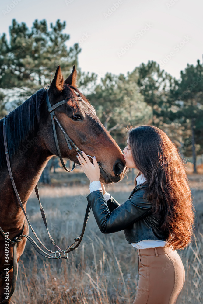 Obraz premium Beautiful elegance woman kisses your hokse. Has smiling face, brown leather jacket and hat. Has slim body. Portrait nature. People and animals. Equestrian. Close up.