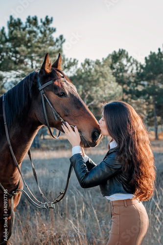Beautiful elegance woman kisses your hokse. Has smiling face, brown leather jacket and hat. Has slim body. Portrait nature. People and animals. Equestrian. Close up. 