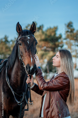 Beautiful elegance woman kisses your hokse. Has smiling face, brown leather jacket and hat. Has slim body. Portrait nature. People and animals. Equestrian. Close up.