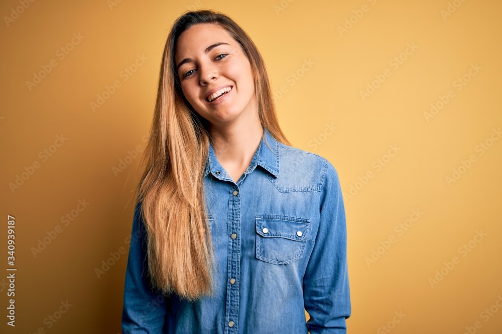 Young beautiful blonde woman with blue eyes wearing denim shirt over yellow background with a happy and cool smile on face. Lucky person. Young beautiful blonde woman with blue eyes wearing denim shirt over yellow background with a happy and cool smile on face. Lucky person.
