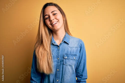 Young beautiful blonde woman with blue eyes wearing denim shirt over yellow background with a happy and cool smile on face. Lucky person.