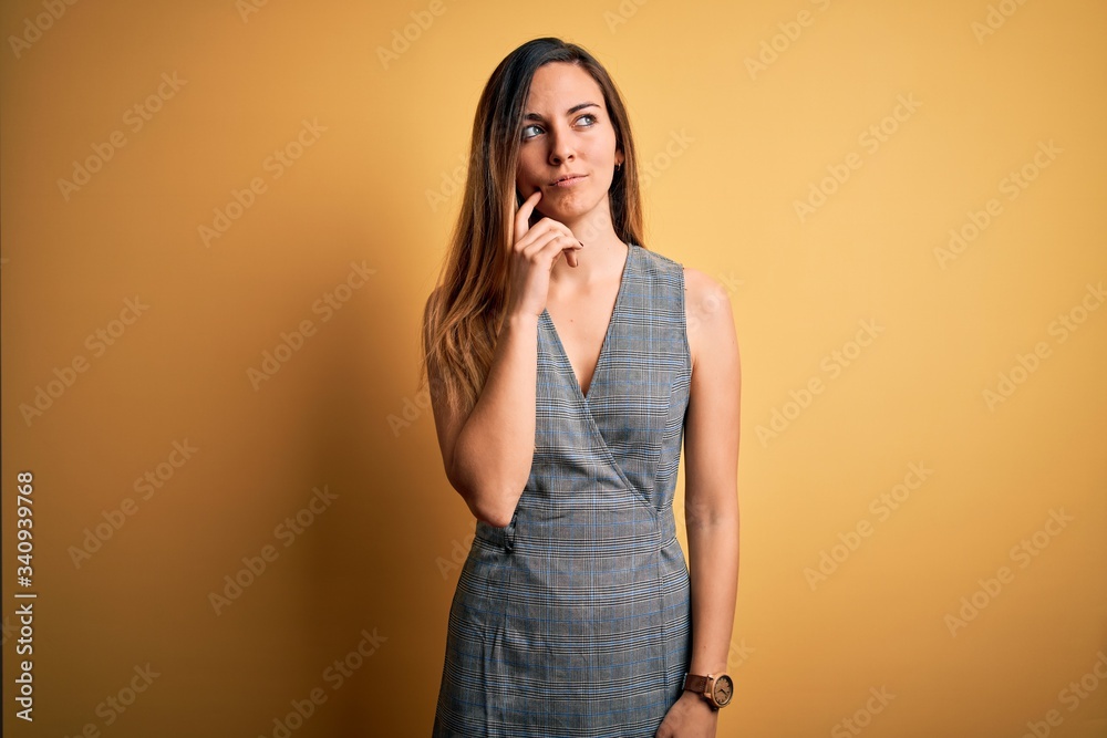 Young beautiful blonde woman with blue eyes wearing dress over yellow background with hand on chin thinking about question, pensive expression. Smiling with thoughtful face. Doubt concept.