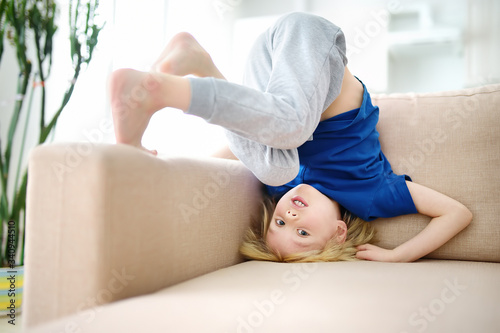 Little boy is standing on own head on sofa at home while coronavirus epidemic. Bored lonely child. Difficulties of family with preschool children during quarantine.