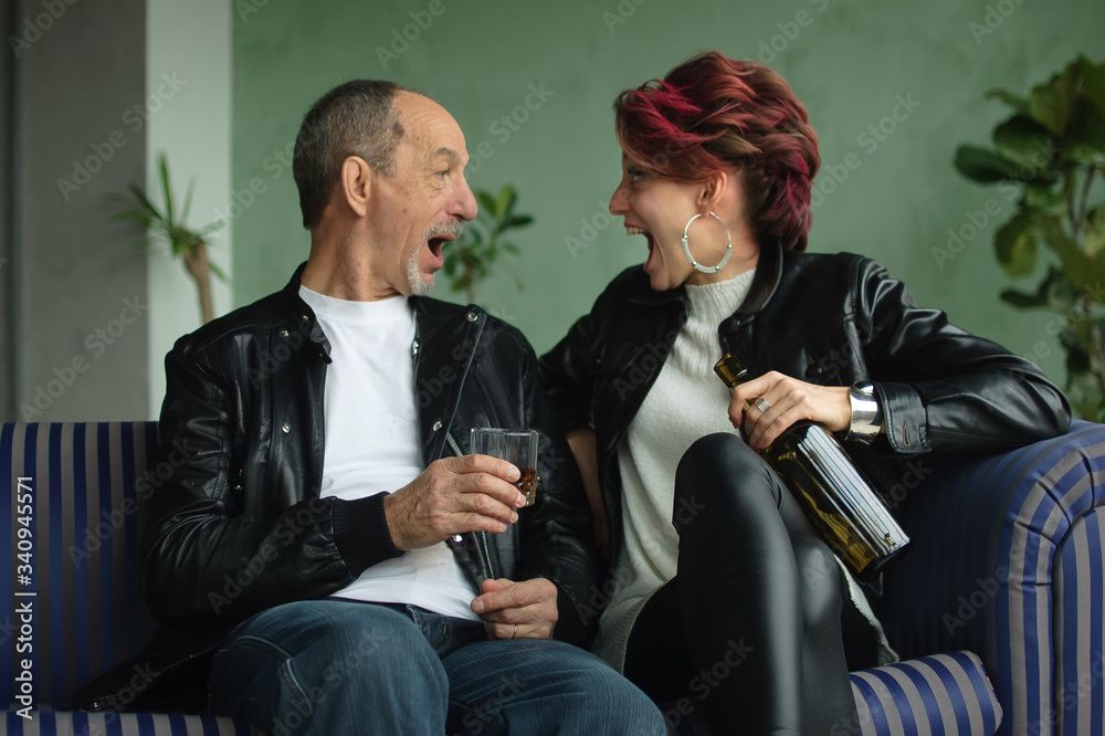 Young wife and senior husband are drinking because of potency problem in their family. Man holds glass of whisky, woman is sitting with bottle in hand, they are screaming to each other