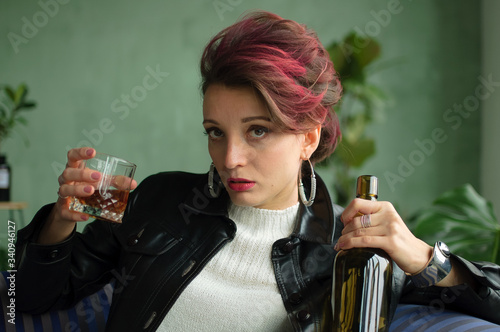 Beautiful sexy woman with dark pink hair in black leather jacket is drinking whisky from a glass sitting on blue sofa at home in loft style room, female alcoholism, alcohol addiction